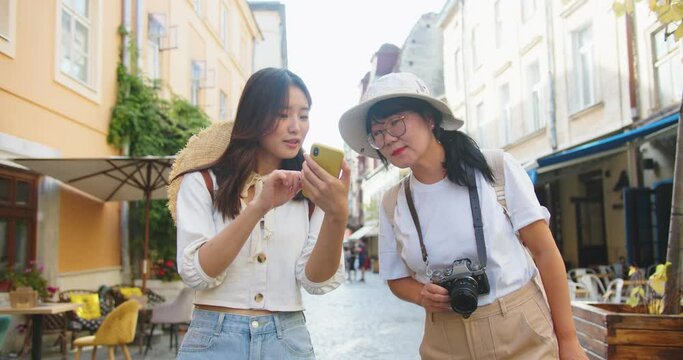 Portrait of happy mother and daughter traveling together. Beautiful woman in hat taking photo on camera outdoors. Young Asian female tourist tapping and browsing on cellphone while walking in city