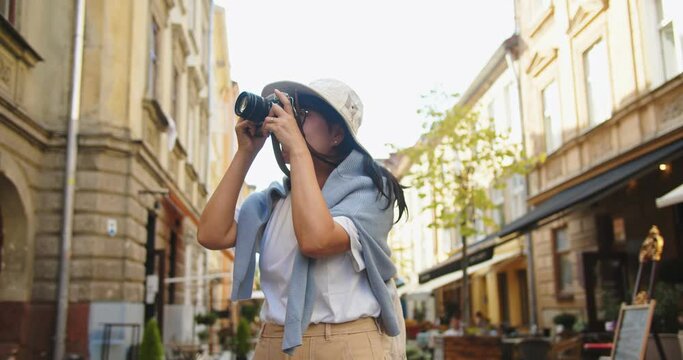 Cheerful Asian beautiful female in hat and glasses taking photo on camera while walking in city on street. Portrait of happy pretty woman takes pictures outdoors while traveling. Urban tourism concept