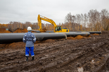 an engineer supervises the laying of the pipeline. In the background, an excavator
