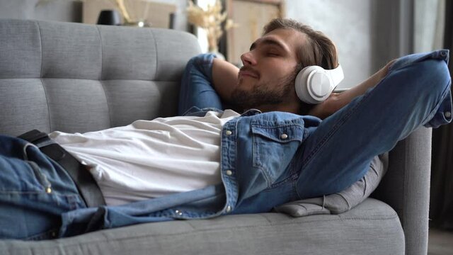 Happy millennial hipster guy wearing modern wireless headphones, choosing enjoying favorite music from mobile application, relaxing alone on comfortable couch in living room.