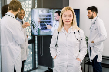 Attractive mature blond woman doctor, posing to camera. Multiracial colleagues near big screen on background, discussing the CT of patient and consulting with Caucasian specialist online