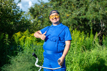 A cheerful man in a blue jumpsuit mows the grass with a trimmer.