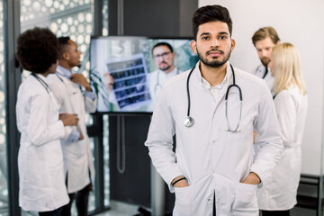 Young handsome Indian male doctor with stethoscope, in modern clinic. International team of doctors on the background, analyzing patient's CT during video conference with Caucasian doctors