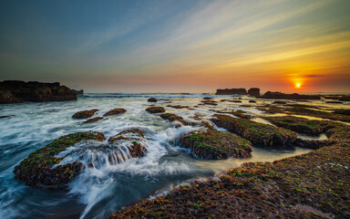 Amazing seascape. Ocean with moving wave. Low tide. Stones covered by green moss and seaweeds. Sun on horizon. Sunset scenery background. Long exposure. Mengening beach, Bali