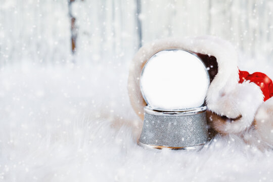 Empty snow globe with copy space surrounded by Santa hat with falling snow. Shallow depth of field with selective focus on snowglobe and free space for text available.