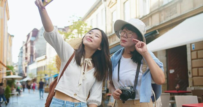 Cheerful Asian mother and daughter making selfie photo on smartphone on street. Portrait of happy female tourists posing and waving hands while taking pictures on cellphone in city. Travel concept