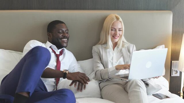 Beautiful multiethnic family spends time on a business trip, watching news on laptop lying on bed in hotel.