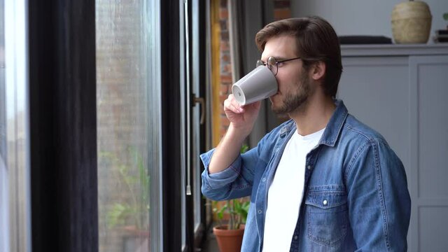 A young man walks up to the window with a mug of coffee or tea and takes a sip