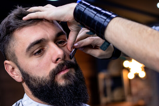 Young bearded man getting haircut by professional at modern barbershop. Beard and sideburns shaving and shaping with vintage straight razor. Movember. Close up. Selective focus. - Powered by Adobe