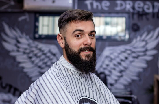 Bearded Man Posing In A Cape After A Haircut And Beard Treatment In Front Of Angel Wings Graffiti On The Wall In A Modern Barbershop. Barber Shop Advertising Concept. Movember. Close Up.