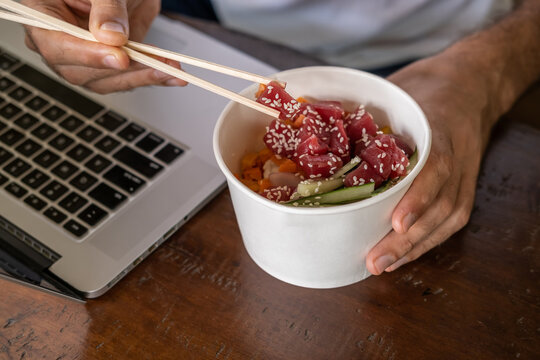 Busy Young Man In Casual Wear Sitting At Desk With A Laptop At Home Or In The Office And Eating Healthy Takeout Poke Bowl Food From Container During Lunch Break. Healthy Lifestyle Concept