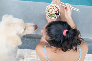 Woman sitting by the pool with her yellow dog and eating takeout poke bowl food