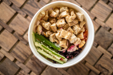 Healthy organic chicken poke bowl with cucumber, rice, carrots, onion served in takeout bowl on wooden table. Healthy lifestyle