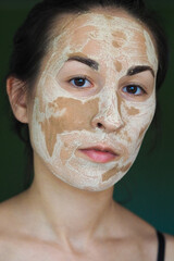  young brunette woman with a clay homemade mask on her face looks into the frame