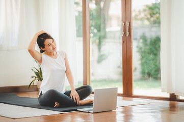 Smiling Asian woman doing yoga neck stretching online class from laptop at home in living room. Self isolation and workout at home during COVID-19.