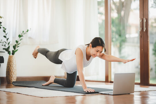 Smiling Asian woman doing one arm one leg plank to exercise core muscle online workout class from laptop at home in living room. Self isolation and workout at home during COVID-19.