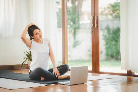 Smiling Asian woman doing yoga neck stretching online class from laptop at home in living room. Self isolation and workout at home during COVID-19.