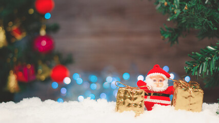 Santa Claus doll with a gift box placed on the snow and the Christmas tree background with bokeh and wood.selective focus. copy space.