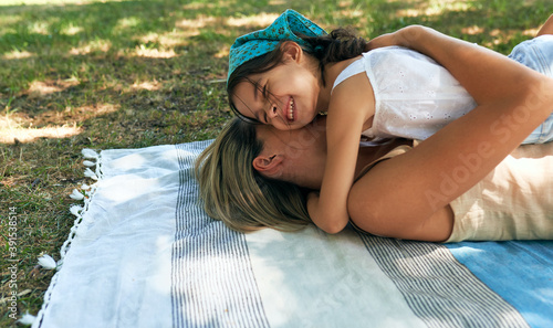 Loving daughter hugging and spending time together with her mother lying on the picnic blanket on the grass in the park. Woman and her child having cozy summer day. Happy Mother's day.