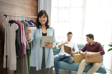 A portrait of a young woman entrepreneur in a clothing shop showing credit card payment with a team. In the background