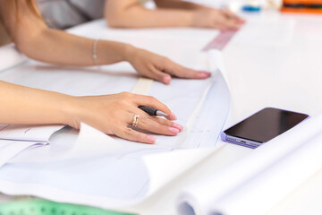 Fashion designer's hands. Closeup of hands drafting a new sewing pattern on paper, working on blueprint plans with a pencil, a ruler, smartphone, tools. Industry, hobby, workspace. Creation process