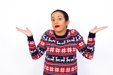 Maybe yes or no. Doubtful Young beautiful Arab woman wearing Christmas sweater shrugs shoulders in...