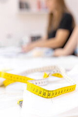 Close up of yellow tailor tape laying on the table with papers in sewing workshop with blurred tailor on background. Sewing accessories, tools. Measuring, centimeter