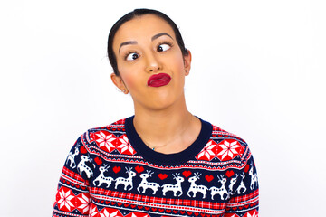 Young beautiful Arab woman wearing Christmas sweater against white wall crosses eyes, puts lips, makes grimace with awkward expression has fun alone, plays fool.