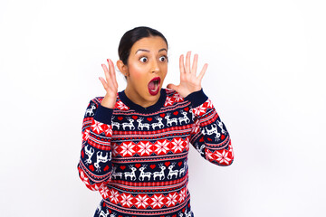 Young beautiful Arab woman wearing Christmas sweater against white wall with shocked facial expression, holding hands near face, screaming and looking sideways at something amazing.