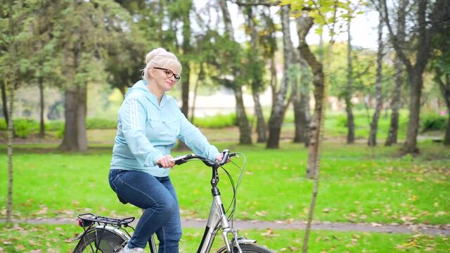 Happy senior woman rides a bicycle in the park and enjoys life. An old female cyclist with a retro bike spends an active healthy life in nature. A walk in the autumn forest or city park outside.