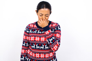 Young beautiful Arab woman wearing Christmas sweater against white wall, holding his nose because...