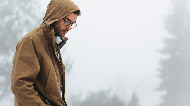 Horizontal outdoor image of a young traveler man walking on a foggy day in mountain. Male wearing raincoat and eyeglasses listening music in headphones outside.