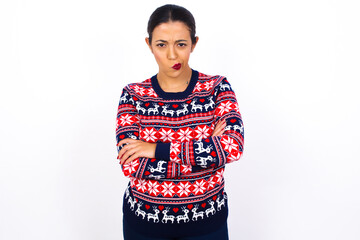 Picture of angry Young beautiful Arab woman wearing Christmas sweater against white wall crossing arms. Looking at camera with disappointed expression.