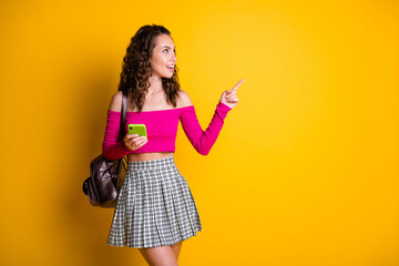 Photo of pretty sweet lady curly hairstyle hold telephone indicate finger look empty space wear bag pink top unclothed shoulders mini skirt isolated yellow color background