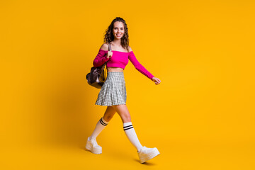 Fototapeta na wymiar Full length photo of pretty sweet lady hold rucksack walk beaming smile wear pink top uncovered shoulders mini skirt stockings sneakers isolated yellow color background