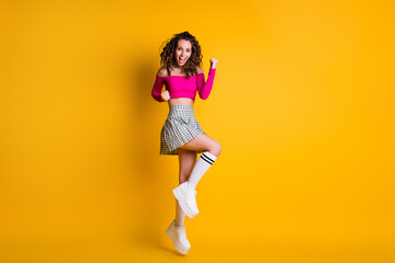 Full body size photo of lovely cute lady wavy hairdo raise arms fists open mouth jump wear pink cropped top uncovered shoulders mini skirt white sneakers isolated yellow color background