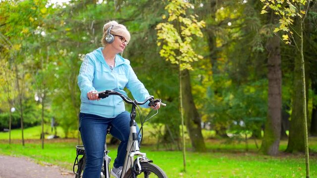 happy senior woman in headphones rides park a bicycle and listens to music audio book. old female cyclist with a retro bike pursues active lifestyle in nature. walk autumn forest or city park outside