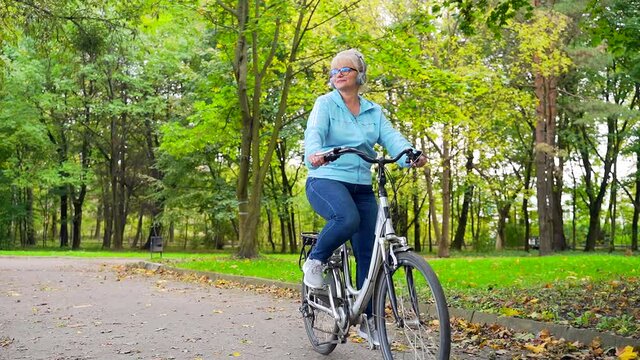 happy senior woman in headphones rides park a bicycle and listens to music audio book. old female cyclist with a retro bike pursues active lifestyle in nature. walk autumn forest or city park outside