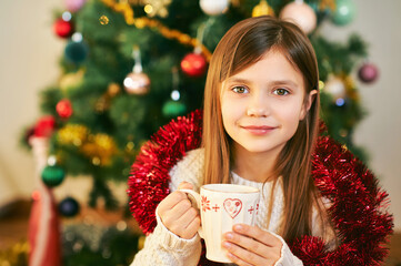 Portrait of sweet little girl sitting next to Christmas tree, wearing white cozy pullover, holding cup with hot chocolate, winter holidays