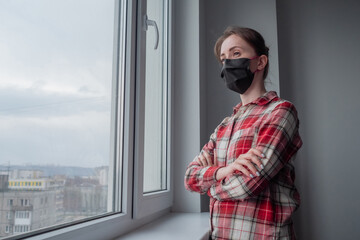 Naklejka premium Portrait of pensive woman wearing black medical face mask, red plaid shirt and looking out of window in room with grey wall at home. Self isolation, quarantine, COVID 19, coronavirus, safety concept
