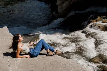 Traveler girl is sitting on the rock near waterfall and looking toward. Travelling in Karpathian mountains. Cascade waterfall. Beautiful landscape