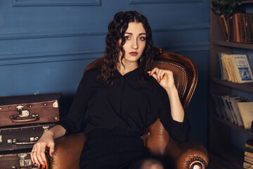 Fototapeta na wymiar Close up portrait of young elegant woman sitting in the luxury room on the old antique armchair, photostudio apartment background. Confident female with curly dark hair looking at the camera.