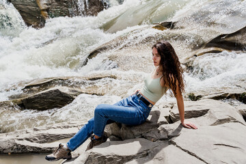 Traveler girl is sitting on the rock near waterfall and looking toward. Travelling in Karpathian mountains. Cascade waterfall. Beautiful landscape