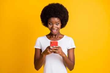 Photo portrait of shocked astonished african american woman holding phone in both hands wearing white t-shirt isolated on vivid yellow colored background