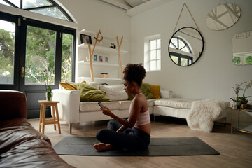Young african woman using cellphone while sitting on yoga mat at home