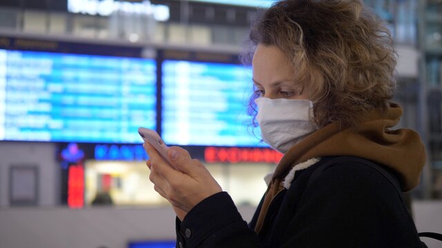 Public transport sector during coronavirus outbreak. Face masked woman checking schedule board with boarding pass on phone