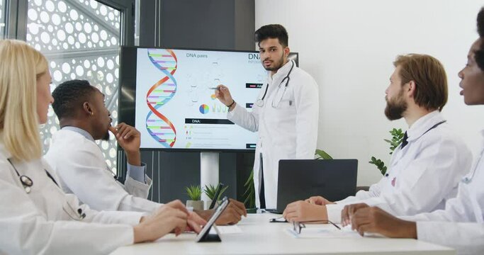 Front view of good-looking confident experienced bearded medical worker holding a presentation on interactive screen for cvalified diverse male and female colleagues