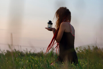 Young smiling beautiful woman with red dreadlocks holding a lantern in sitting in meadow on grass