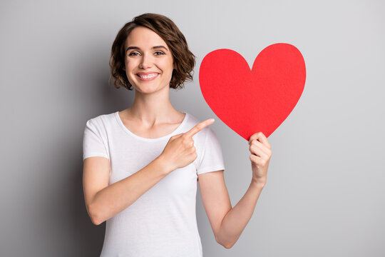 Photo of young happy smile girl point index finger hold big heart valentine day advertise offer isolated over grey color background