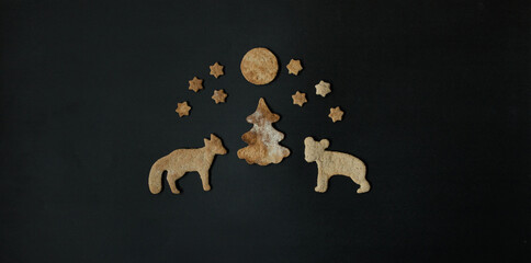 Christmas holidays spirit concept: gingerbread or season biscuits shaped as a fir tree with forest animals under stars and moon on black. Cute greetings card. Creative flat lay. Top view, copy space.